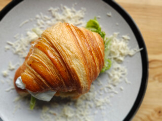 Close-up photography of a crispy croissant sandwich filled with lettuce leaf, tomato, and white cheese on a white plate. Wooden table. Top view. Blurred backdrop.