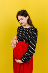 Happy pregnant woman holding a teddy bear against her belly at Colored background. Young mother is...