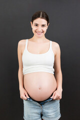 Happy pregnant woman in unzipped jeans showing her naked belly at colorful background with copy...