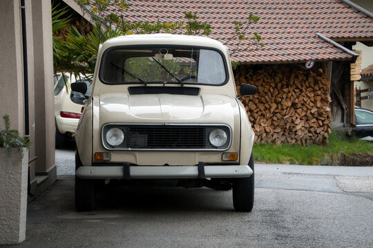 Ein altes, abgemeldetes Auto auf einem Vorplatz an einer Hauswand geparkt, mit einem zweiten Auto und gestapeltem Brennholz im Hintergrund