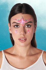 Cropped close-up shot of a woman with colored shimmering crystals on her face. A beautiful young woman in a white dress with makeup and jewel stickers on her forehead and cheeks is posing on the blue 