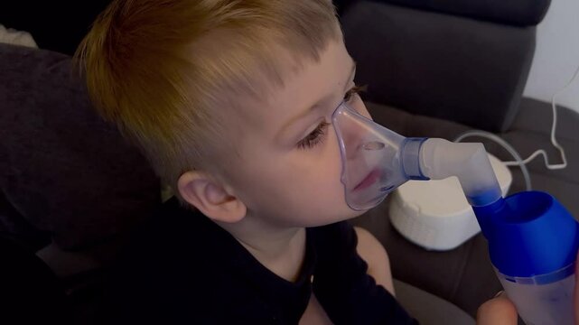 Close up of little caucasian sick boy holding a inhalation mask on his face to breath saline solution, at home