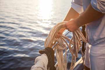 Man sailing on yacht on sea at sunset. Close-up of sailor hands that moor, tie up rope. Traveler enjoying adventure in summer vacation. Lifestyle moment on ecological transport in sustainable travel