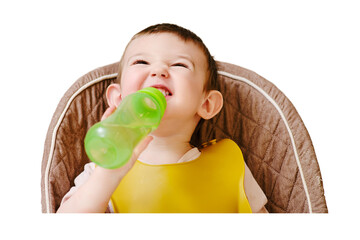 Happy toddler baby drinking from a bottle while sitting on a high chair, isolated on white background. The child boy in the bib has lunch in the home kitchen. Kid aged one year six months