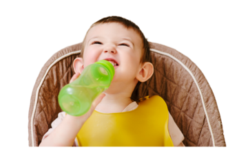 Happy toddler baby drinking from a bottle while sitting on a high chair, isolated on white background. The child boy in the bib has lunch in the home kitchen. Kid aged one year six months