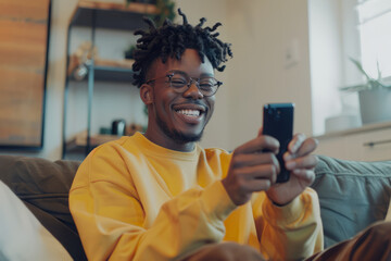 Young Afro-American man sits on a sofa at home, holding a smartphone. His radiant expression...