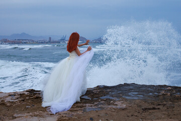 Mujer pelirroja con vestido de novia en la playa y salpicaduras de ola de mar con gesto de parar el...