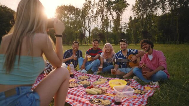 Group of multi-ethnic teenagers toast and celebrate during a picnic in the park. Millennials sing songs while playing the guitar and a girl capturing the moment with a vintage camera.