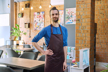 Young man worker, owner in apron looking at camera in restaurant, coffee shop