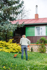 Lawn care in yard around wooden cottage. Woman cutting grass by string trimmer. Spring gardening