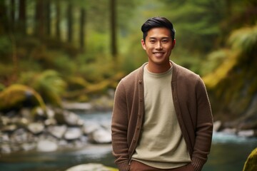 Portrait of a grinning asian man in his 20s dressed in a warm wool sweater isolated in tranquil forest stream