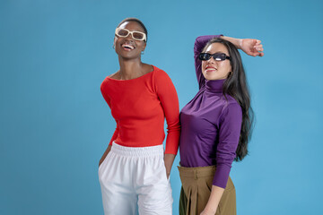 Two female fashion models with sunglasses posing smiling happy on a blue studio background. High quality photo