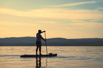 Silhouette of a man paddling on a stand up paddle boarder or SUP on the lake at sunset. Hills in...