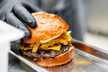 Person in black gloves holding a freshly made burger with a glossy bun, melted cheese, and various toppings, highlighting the appeal of fast food