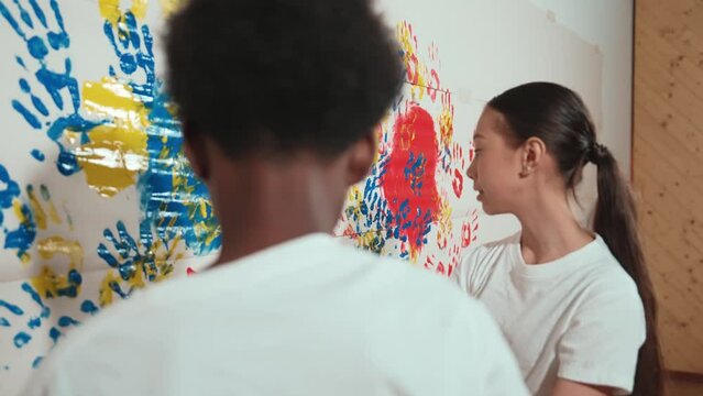 Asian girl use hand to stamp the wall with red color in art lesson. Multicultural high school student wear white shirt while paint room with colorful hand print. Creative activity concept. Edification