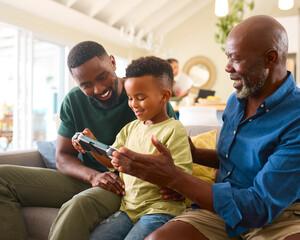 Grandfather With Father And Grandson Play Handheld Computer Game At Home With Family In Background