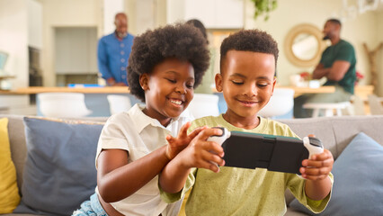 Boy And Girl  Playing With Handheld Gaming Device At Home With Multi Generation Family In Background