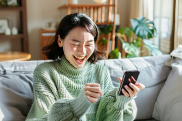 With excitement, a Japanese woman, holds a smartphone while sitting on a sofa at home. Her delighted expression indicates satisfaction as she looks at the mobile smartphone screen, gesturing yes