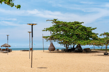 On the yellow sand of the beach, in the shade of a large tree, there are sun loungers and other beach furniture. 
