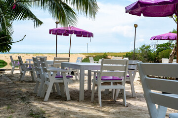 On the shore of a tropical sea on a sandy beach in the shade of trees there are tables and chairs of a street cafe.