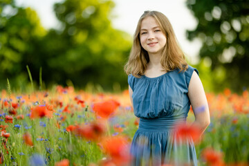 Beautiful teenage girl admiring poppy and knapweed flowers in blossoming poppy field on sunny summer day. - obrazy, fototapety, plakaty