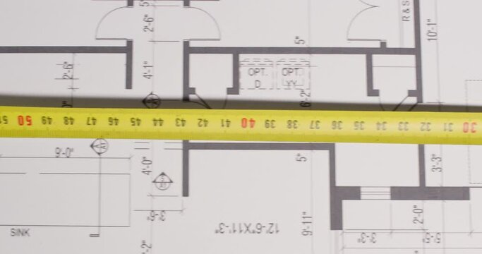 A builder moves a tape measure with his hand along a paper drawing of a building, pulls out a ruler to measure, close-up.