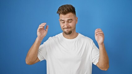 A young bearded man in a white shirt stands against a blue wall outdoors, covering his ears with a frustrated expression.