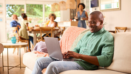 Parents Indoors At Home On Sofa Looking At Laptop With Grandparents And Children In Background