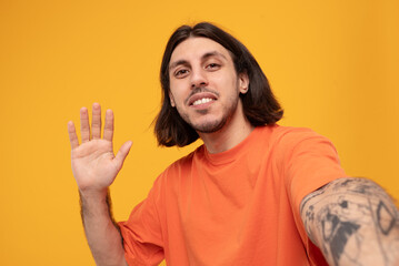 Portrait of cheerful middle eastern man wearing t-shirt greeting and taking selfie photo while standing isolated on orange background in studio