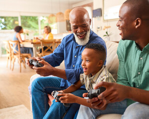 Grandfather With Father And Grandson Playing Computer Game At Home With Family In Background