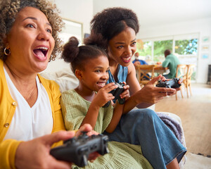 Grandmother With Mother And Granddaughter Playing Computer Game At Home With Family In Background