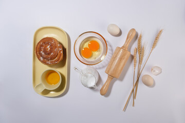 Flat lay baking ingredient, utensil on minimalist white kitchen counter top, cinnamon roll, a cup of oil, cracked egg in transparent bowl, water and rolling pin. Food background with space for text