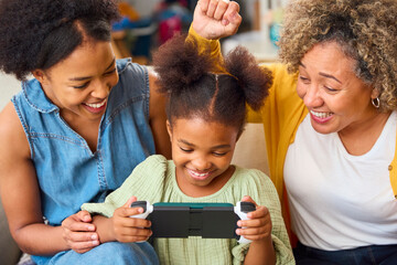 Grandmother With Mother And Granddaughter Playing With Handheld Gaming Device At Home With Family