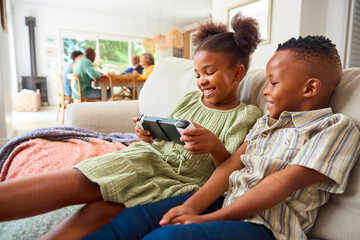 Boy And Girl Playing With Handheld Gaming Device At Home With Multi-Generation Family In Background