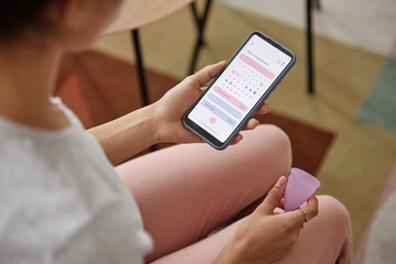 Closeup of young woman holding menstrual cup and smartphone with period tracker app on screen copy space