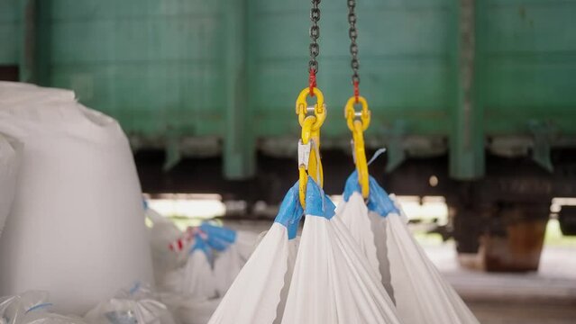 Industrial crane hooks lifting heavy white bags in a warehouse. Factory rigging working processes, moving materials with cathead inside workshop. Loading bags into train wagons