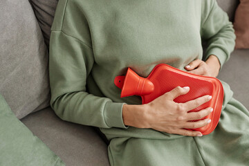 Close up of unrecognizable woman holding red hot water bottle to stomach suffering from period...