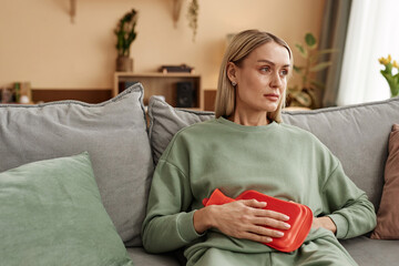 Portrait of adult woman holding red hot water bottle to stomach suffering from period cramps sitting on couch at home copy space