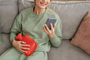 Close up of smiling woman holding red hot water bottle to stomach and using smartphone at home copy space