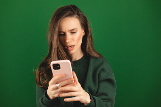 Mad and shock young brunette beautiful woman using mobile phone isolated on green background. Yelling unhappy woman hold mobile phone. Disappointed sad upset lady horrified impressed news. WTF. Oh no.