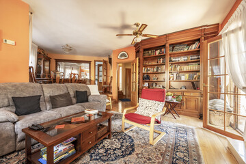 Two-story living room in a house with wooden furniture, a bookcase filled with many books, large rugs and varnished oak hardwood floors