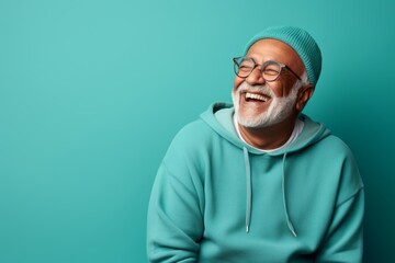 Obraz premium Portrait of a smiling indian man in his 80s sporting a comfortable hoodie in front of soft teal background