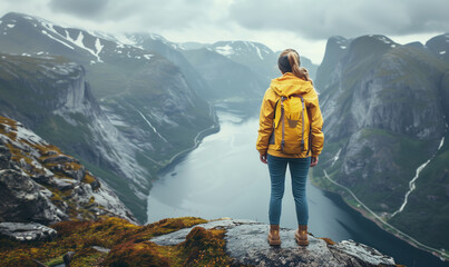 Portrait of a woman in a yellow raincoat standing from her back on a troll's tongue high in the mountains of Norway against the background of fjords
