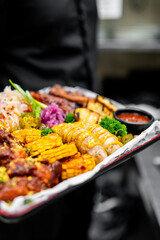 Person holding a tray with an assortment of appetizing food including grilled corn, sausages, and sauces, showcasing a vibrant and varied meal