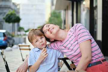 Young mother and her son having sweets in outdoor restaurant on summer day. Sitting together, hugging and laughing, enjoying weekend. High quality photo