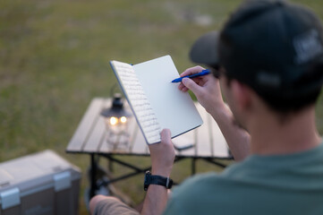 Man seated in a folding chair writing in a notebook with camping gear, enjoying the tranquility of...
