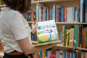 A young mother wearing a white tank top is flipping through a children's book in the bookstore