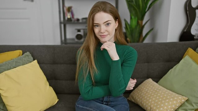 Cheerful young blonde woman oozing confidence, wearing sweater at home, arms crossed, hand on chin, beaming into camera, lost in positive thoughts.