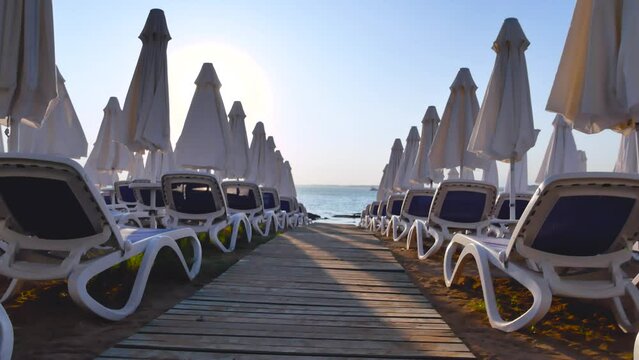 Lots of sun loungers in the rays of the setting sun, lowered sunshades, a wooden path leads to the sea. Evening on the sea beach.