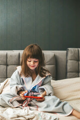 A cute little girl wearing a bathrobe sitting on a bed, completely engrossed in her smartphone. Concept: kids and smartphones